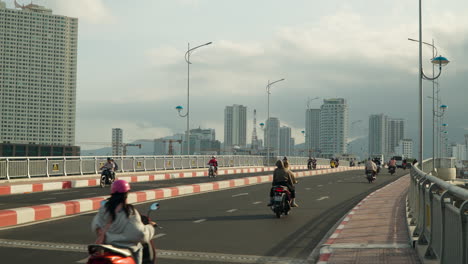 Vietnamese-People-Riding-Motorcycles-Crossing-Xom-Bong-Bridge-in-Nha-Trang-City,-Skyscraper-Buildings-in-Background,-Vietnam---in-slowmo