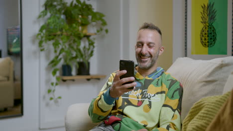 Kaukasischer-Mann-Sitzt-Auf-Der-Couch-Und-Lacht,-Während-Er-Lustige-Videos-Auf-Dem-Mobiltelefon-Ansieht