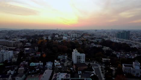 Cityscape-at-sunset-with-vivid-skies-and-urban-silhouette,-hints-of-autumn-in-tree-colors,-aerial-view