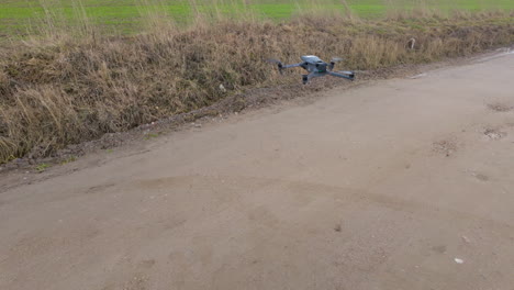A-drone-with-a-grey-body-and-blue-lights-is-flying-low-over-a-dirt-road,-with-dried-grass-and-a-slope-on-the-side