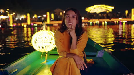 Attractive-Vietnamese-woman-on-river-boat-at-night-looking-at-light-reflections,-Hoi-An,-Vietnam