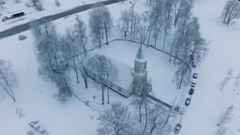 Snow-covered-church-in-Renda-village-with-surrounding-bare-trees,-aerial-view,-moving-forward,-tilt-down