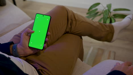 Detail-of-a-male-hand-using-a-cell-phone-sitting-on-the-sofa-while-relaxing---cell-phone-screen-in-green-chroma-key-for-graphic-insertion
