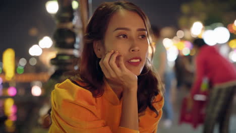 Mujer-Vestida-De-Amarillo-Ao-Dai-Luciendo-Pensativa-Por-La-Noche-En-Hoi-An,-Vibrantes-Luces-De-La-Calle-En-El-Fondo,-Primer-Plano