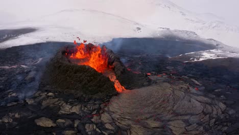 Un-Dron-De-4k,-Cinemático-Aéreo,-Tomas-únicas-De-Un-Pico-Volcánico-En-Erupción-Derrama-Lava-Rápidamente-Y-Campos-Nevados-E-Invernales-Al-Fondo-En-Islandia