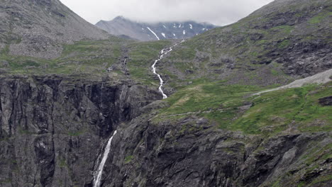 Aerial-dolly-shot,-pushing-in-slowly-on-a-long,-thin-river,-winding-down-a-steep,-green,-mossy-hillside-by-the-Trollstigen,-Norway