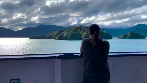 Dark-haired-woman-in-sweatshirt-leans-on-boat-railing-and-looks-out-at-islands-and-cloudy-skies-in-Queen-Charlotte-Sound-in-New-Zealand