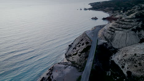 Aphrodite's-Rock-Paphos---Coastal-road-curving-along-a-cliffside,-sunset,-ocean-view,-aerial-perspective
