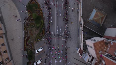 Vista-Aérea-De-Niños-Bailando-En-La-Calle-Con-Trajes-Tradicionales-Con-Banderas-Ondeando,-Festival-De-Folklore-Infantil-Licidersko-Srce-En-Uzice,-Serbia