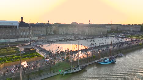 People-gathering-in-Place-de-la-Bourse-plaza-square-during-Wine-Fair-with-sailing-ships-at-dock,-Aerial-dolly-right-shot