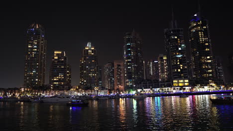 Dubai-Marina-at-Night,-Lights-on-Skyscrapers,-Touristic-Boats-and-Reflection-on-Water