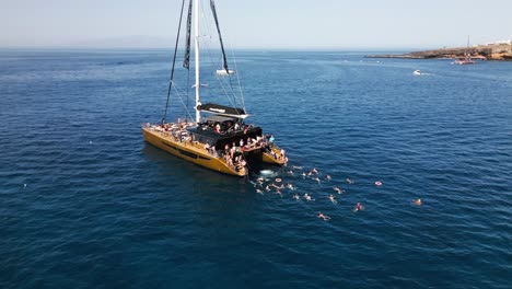 El-Hombre-Salta-Del-Barco-Al-Agua,-Catamarán-En-Agua-De-Mar-Azul,-Hermosa-Bahía-En-Una-Isla,-Tenerife,-Barco,-Verano,-Drone