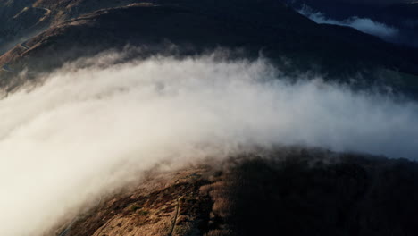 Densas-Nubes-Rodando-Sobre-Terreno-Montañoso-Al-Atardecer,-Vista-Aérea