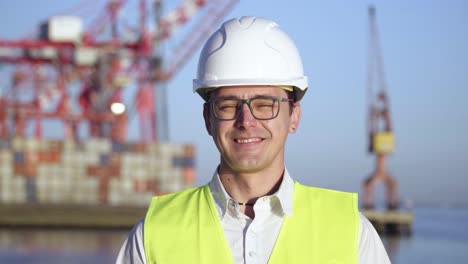 Retrato-De-Un-Joven-Y-Alegre-Inspector-De-Control-Con-Gafas-Y-Un-Casco-Blanco-Parado-Frente-Al-Puerto-De-Carga-De-Contenedores