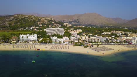 Drone-panning-from-the-right-to-the-left-side-of-the-frame,-showing-the-Playa-del-Port-Pollenca-beach-resort,-in-Malloraca,-Spain,-with-the-mountains-and-luxurious-hotels-in-the-background