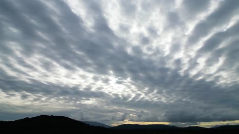 Luftaufnahme-Mit-Kameraschwenk,-Die-Den-Sonnenuntergangshimmel-Voller-Hoher-Graublauer-Wolken-Und-Orangefarbener-Farben-Des-Sonnenuntergangs-über-Der-Silhouette-Der-Berge-In-Blau-Und-Schwarz-Aufnimmt