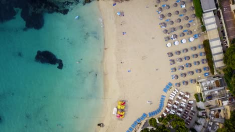 Toma-En-órbita-Y-Descendente-Del-Complejo-Playero-De-Playa-Lletes,-Que-Muestra-Las-Sombrillas-Alineadas-En-Filas-Donde-Los-Bañistas-Se-Tumban-Para-Tomar-El-Sol-En-La-Isla-Mediterránea-De-Mallorca-En-España.