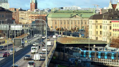 Subway-train-drives-under-trafficked-bridge-in-sunny-Stockholm,-Sweden