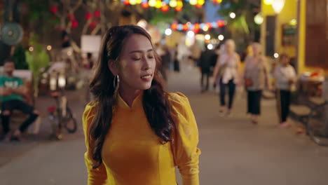 Woman-in-vibrant-yellow-Ao-Dai-strolling-through-Hoi-An's-lively-evening-market,-lanterns-glowing