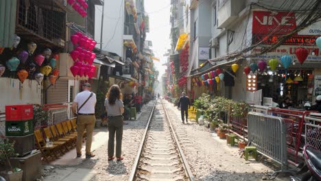 Couple-explores-vibrant-Train-Street-in-Hanoi-with-shops-and-colorful-lanterns,-sunny-day