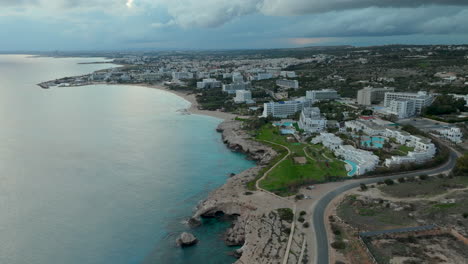 panoramic-aerial-view-of-Ayia-Napa,-expanse-of-the-azure-Mediterranean-Sea-alongside-a-curved-coastline-dotted-with-hotels-and-resorts,-town-stretching-into-the-interior-landscape-under-a-moody-sky