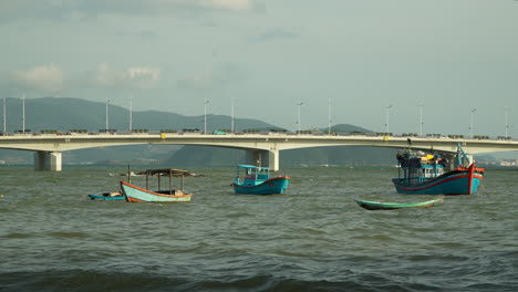 Barcos-De-Pesca-De-Madera-Anclan-En-El-Agua-Del-Río-Cai-En-La-Costa-De-La-Aldea-De-Cu-Lao-Con-El-Puente-Tran-Phu-Y-Las-Montañas-En-El-Fondo,-Nha-Trang-Vietnam