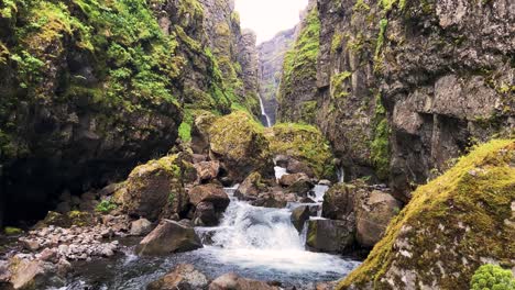 Moss-covered-cliffs-with-a-waterfall-in-a-lush-Icelandic-canyon,-cloudy-daylight