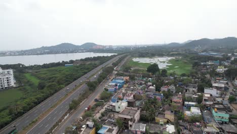 Drone-Shot-Of-Highway-Near-A-Lake-Surrounded-By-Buildings-And-Hills-in-The-Background