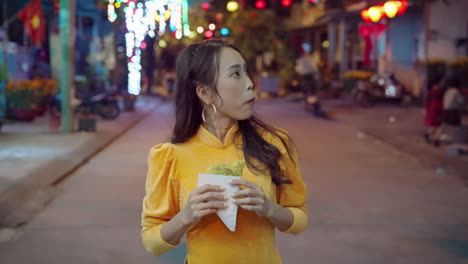 Slow-motion-of-Vietnamese-woman-in-traditional-clothing-walking-and-eating-local-pizza-in-Hoi-An-street-at-night