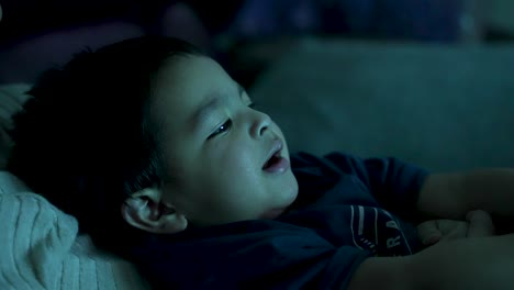 British-Asian-18-month-old-relaxes-on-the-sofa,-engrossed-in-watching-TV,-with-gentle-light-reflections-casting-a-cozy-atmosphere,-embodying-the-essence-of-leisurely-home-free-time