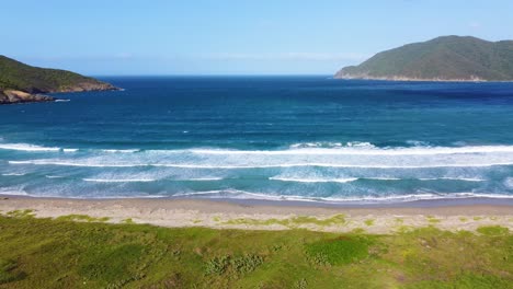 A-vibrant-beach-with-waves-crashing,-surrounded-by-greenery-and-hills,-under-a-clear-sky,-aerial-view