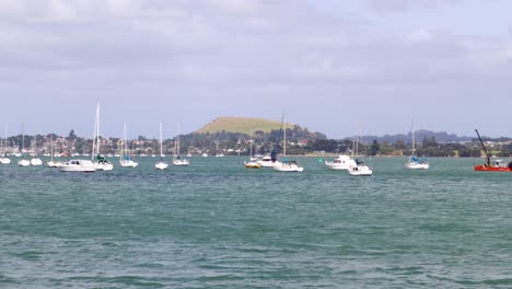 A-handheld-shot-of-a-group-of-boats-floating-on-a-lake-with-Mount-Wellington-volcano-in-the-background-in-Auckland,-New-Zealand,-on-a-windy-and-cloudy-day