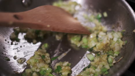 Close-up-wooden-spatula-stirring-and-turning-diced-onions,-garlic,-and-shallots-as-they-sizzle-and-soften