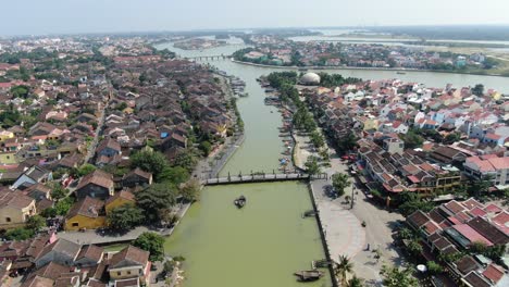 Drohnen-Luftaufnahme-In-Vietnam,-Die-An-Einem-Sonnigen-Tag-über-Den-Braunen-Flusskanal-Von-Hoi-An-In-Der-Stadt-Fliegt,-Kleine-Backsteinhäuser-Und-Holzboote