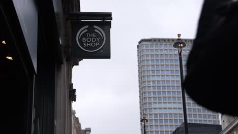 In-slow-motion-a-person-walks-past-The-Body-Shop-store-sign-on-Oxford-Street-with-the-Centerpoint-tower-block-in-the-background