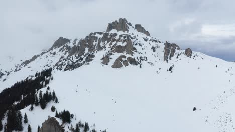 Montañas-Ciucas-Cubiertas-De-Nieve-Bajo-El-Cielo-Nublado,-Pinos-En-Primer-Plano,-Vista-Aérea