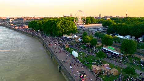 Orilla-Del-Río-Garona-Con-Noria-Llena-De-Multitudes-Durante-La-Feria-Del-Vino,-Toma-Aérea