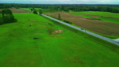 Luftdrohnenlandschaft-Auf-Nasser,-Grüner,-Abgestorbener-Grasstraße-Durch-Landwirtschaftliche-Felder-Auf-Dem-Land,-Auto-Fährt-Langsam-Mit-Feuchter,-Tageslichtblauer-Skyline,-Dorfrand