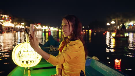 Pretty-Vietnamese-woman-in-traditional-yellow-dress-takes-phone-photos-on-a-night-boat-in-Hoi-An,-Vietnam