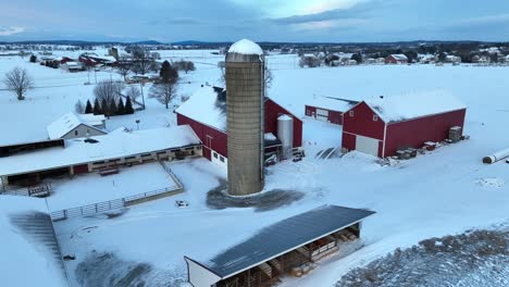 Snowy-farm-with-red-barns,-a-silo,-and-bare-trees-under-a-cloudy-sky