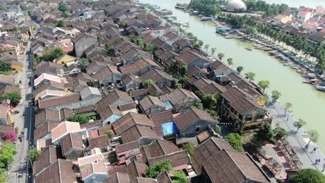 Drone-aerial-view-in-Vietnam-flying-over-Hoi-An-brown-color-river-canal-in-the-city,-small-brick-houses-and-wooden-boats-on-a-sunny-day