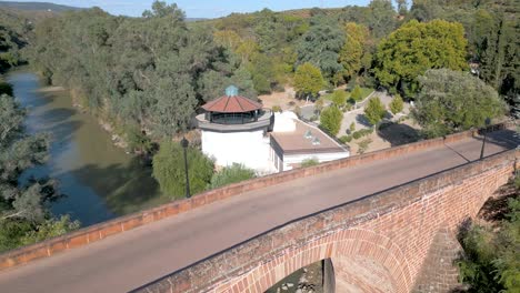 Aerial-of-the-16th-century-bridge-spanning-the-Guadalquivir-River,-traversing-the-buildings-and-gardens-of-Marmolejo-Spa,-situated-within-Jaén-province