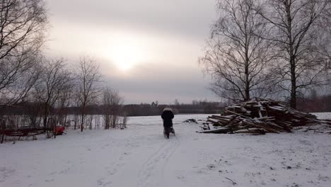 Mother-push-baby-carriage-on-snowy-countryside-yard-near-wood-pile