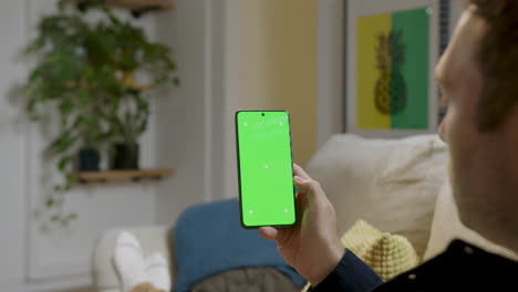 Mann-Hält-Smartphone-Mit-Grünem-Bildschirm,-Berührt-Es,-Als-Würde-Er-Es-Benutzen,-Und-Sitzt-Bequem-Zu-Hause