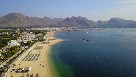 Approaching-drone-shot-showing-the-whole-stretch-of-the-Playa-del-Port-de-Pollenca,-a-popular-tourist-destination-in-the-island-of-Mallorca,-in-Spain