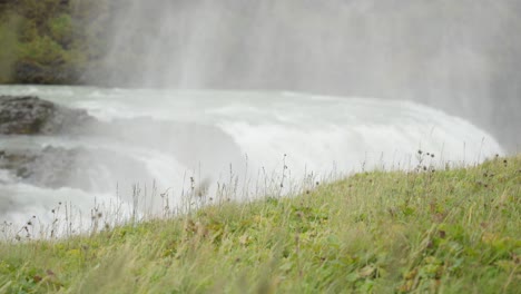 Fernaufnahme-Von-Grünem-Gras,-Das-Auf-Einem-Feld-Weht,-Mit-Einem-Majestätischen-Wasserfall-Im-Hintergrund