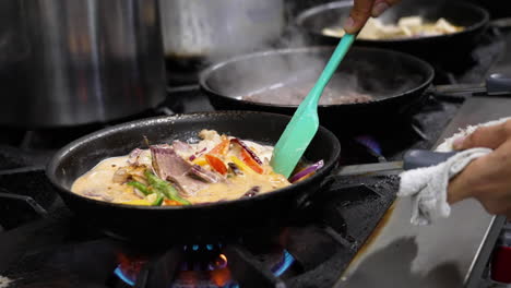 Cinematic-shot-of-crowded-professional-restaurant-kitchen-stove,-line-of-sizzling-skillets-on-range-leading-to-cook-stirring-vegetables-in-sauce,-slow-motion-close-up-slider-4K