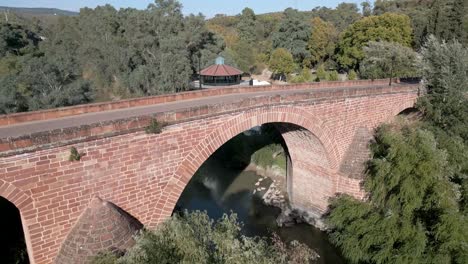 Aerial-of-the-16th-century-bridge-over-the-Guadalquivir-River,-crossing-over-buildings-and-gardens-of-the-Marmolejo-Spa,-in-the-province-of-Jaén