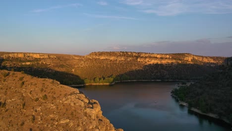 Vuelo-De-Drones-Sobre-Un-Embalse-Que-Se-Encuentra-En-Un-Cañón-De-Piedra-Caliza-Haciendo-Que-Una-Cámara-Gire-A-La-Izquierda-Con-árboles-En-El-Fondo-En-La-Hora-Dorada-Del-Atardecer-Con-Un-Cielo-Azul-En-Verano-Segovia-España