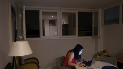 A-woman-uses-a-laptop-indoors-while-a-thunderstorm-is-taking-place-outside,-with-flashes-visible-through-the-home-windows